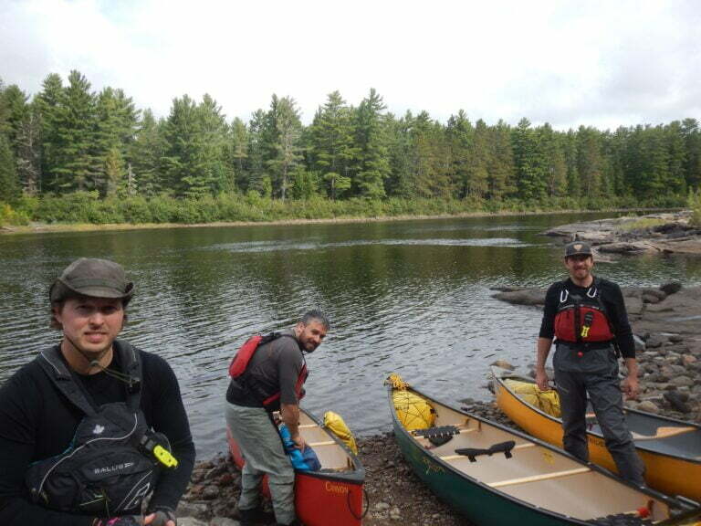 Three men wearing lifejackets posing in front of a river lined with trees while loading canoes with equipment