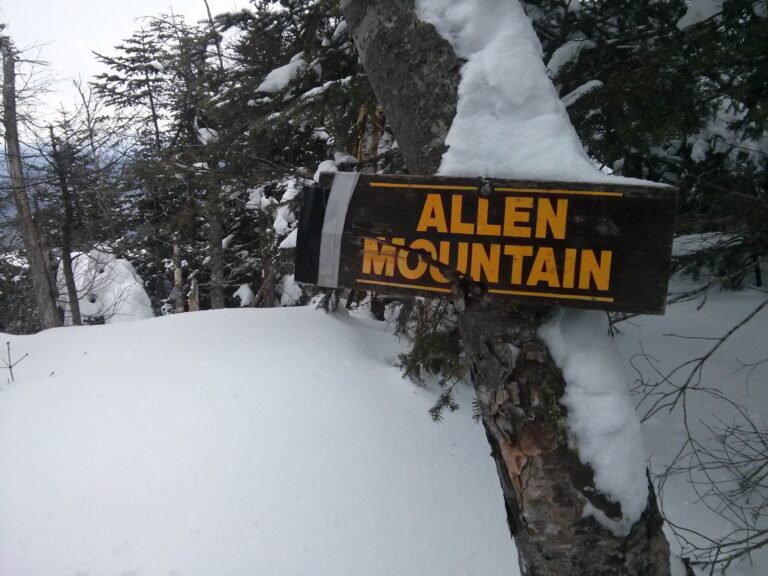 Sign indicating the top of Allen Mountain attached to a tree with snow covering. The sign is broken and held together with tape.