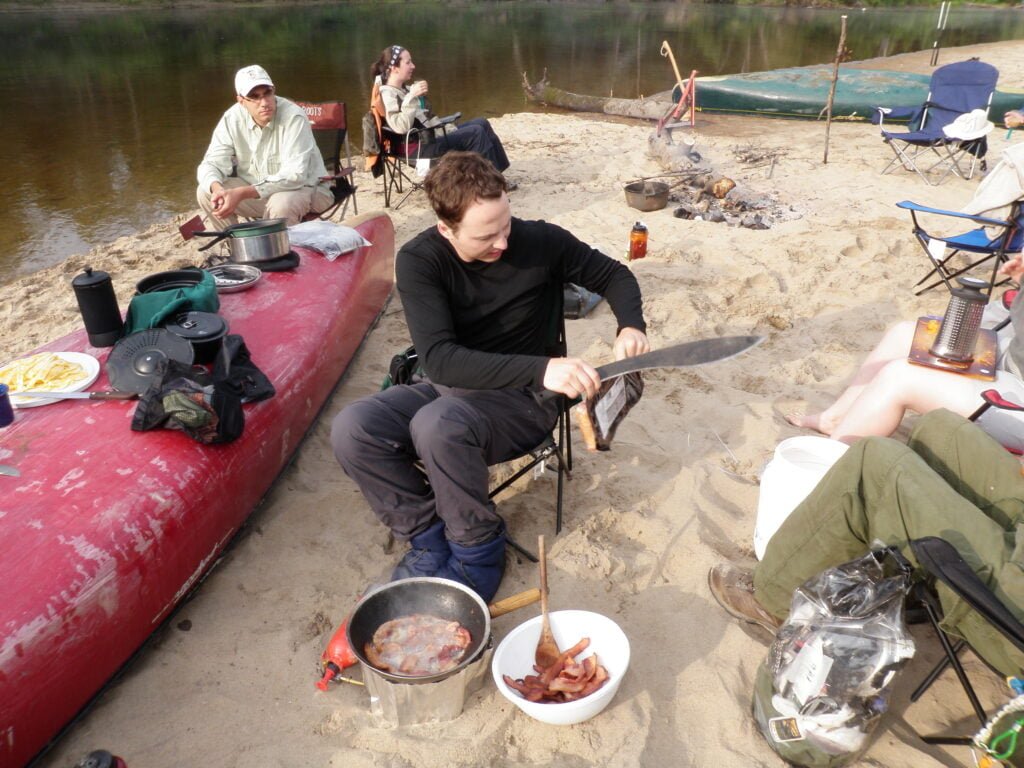 Man opening bacon with machete while sitting down in camping chair in the sand next to a canoe