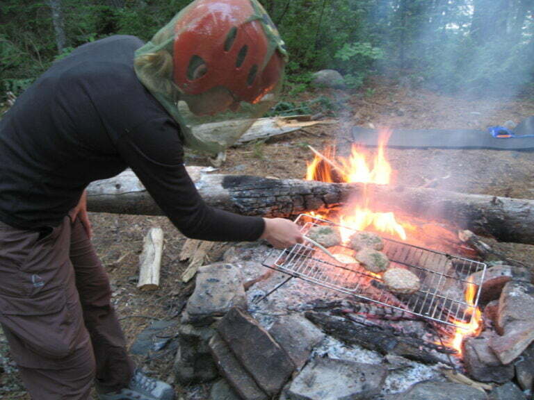 Val cooking hamburgers on the campfire while wearing whitewater helmet and bug net