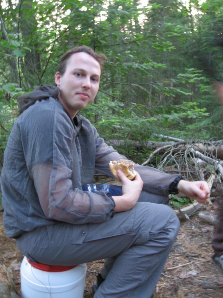 Sunshine (person) sitting on a barrel eating a hamburger while wearing a bug shirt