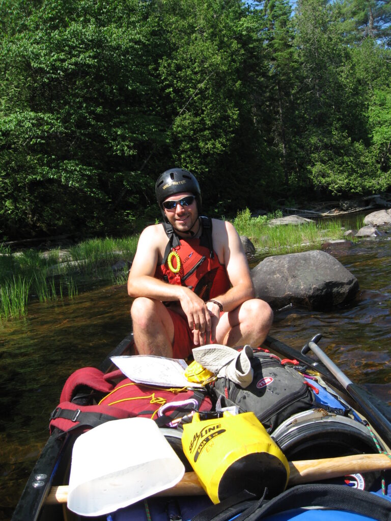 Shirtless man wearing sunglasses lifejacket and whitewater helmet sitting in a canoe filled with waterproofed camping equipment