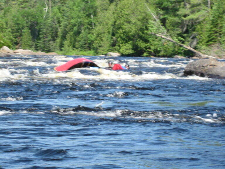 capsized canoe in middle of whitewater rapid with head of paddlers visible above the water line