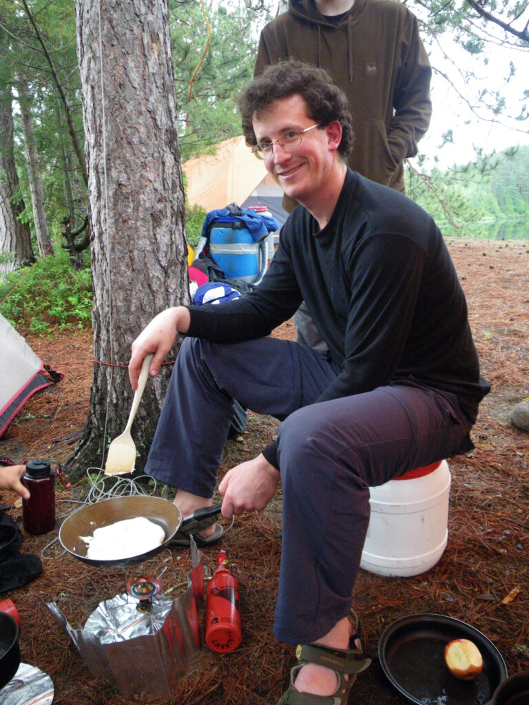 Seb (person), sitting on small plastic food barrel cooking pancakes on camp stove