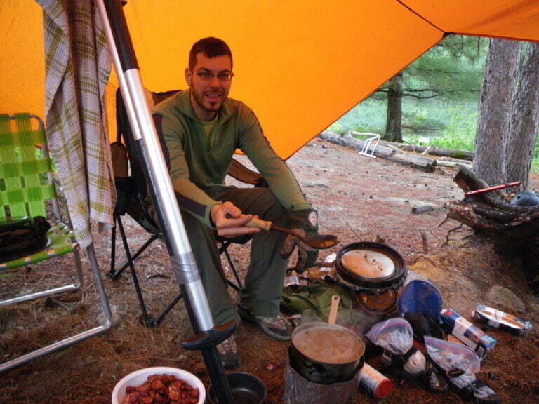 Wissell (person), sitting on a camp chair, cooking noodles on camp stove sheltered by guide tarp