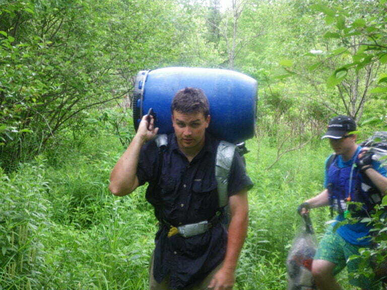 Man carrying two blue waterproof barrels, one on backpack and one on top during portage
