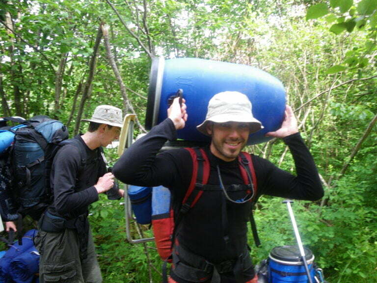 two men portaging blue barrels in the forest