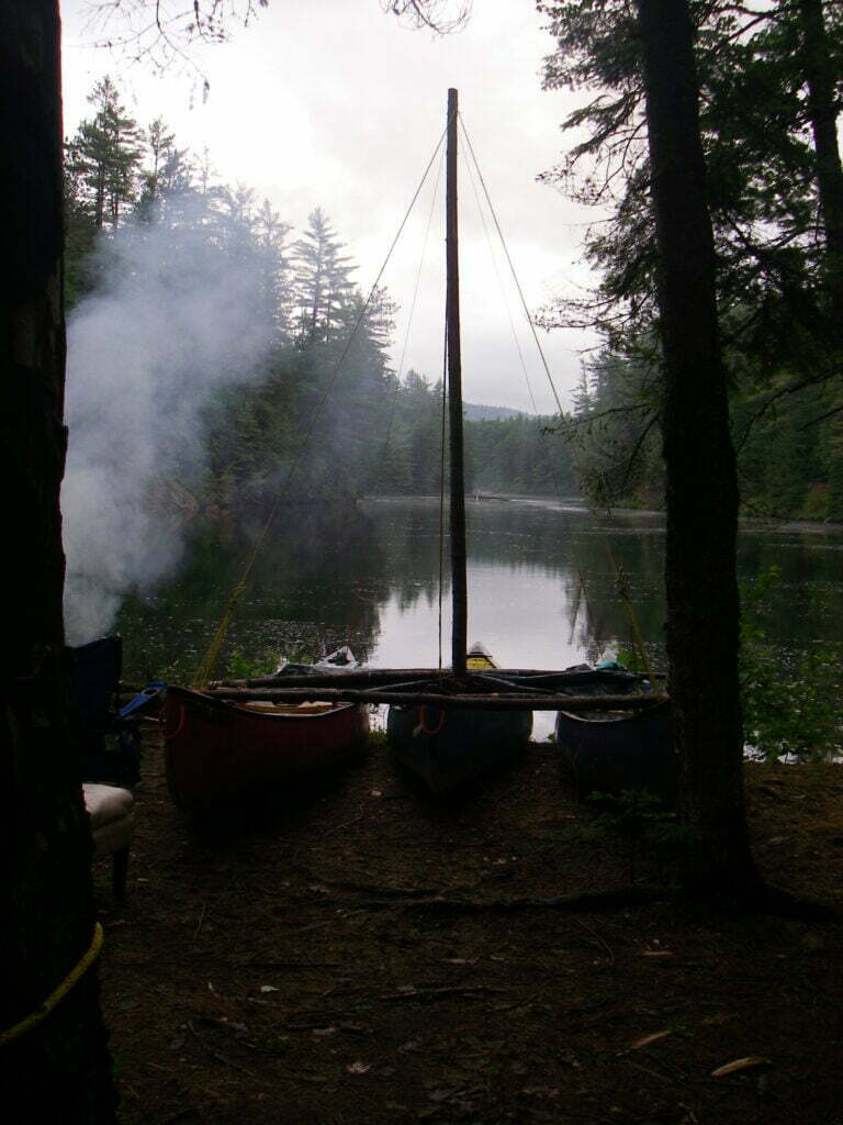 Three canoes lashed together with tree trunks with mast on side of river