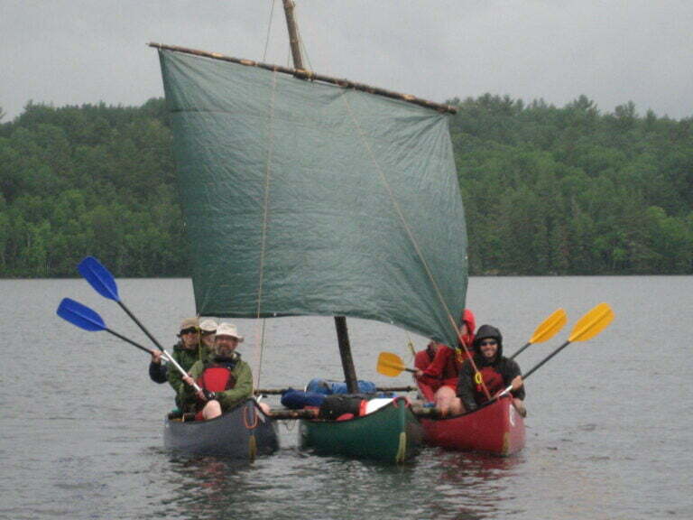 canoe trimaran with sail deployed and paddlers showing paddles outside of water